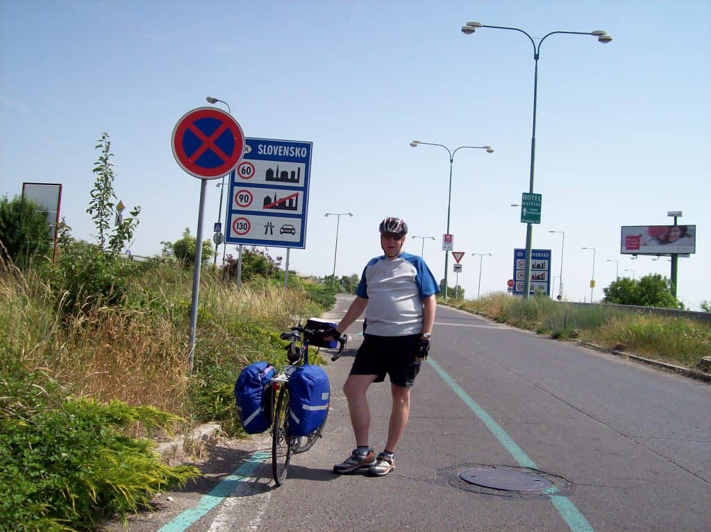 A cyclist stands beside his bike in a designated bike lane at the border between Austria and Slovakia, marked by a road sign displaying the word 'Slovensko' and speed limits. The cyclist, dressed in a white and blue outfit and helmet, pauses near various road signs under a clear blue sky, with dry grass and lamp posts lining the road.