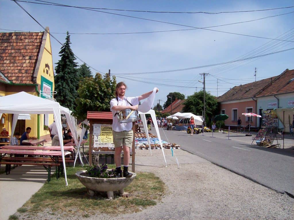 A vibrant street market scene in Raika with a young man standing on a planter, playfully pointing to something out of frame. The street is lined with colorful market stalls and umbrellas, small local shops, and people browsing goods under a clear blue sky.