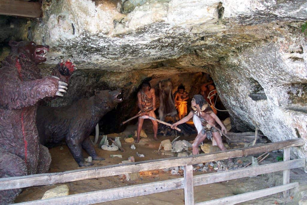 Diorama depicting prehistoric life at La Roque St Christophe in France, with life-sized models of humans in ancient attire and a bear inside a cave dwelling, highlighting the historical significance of the site.
