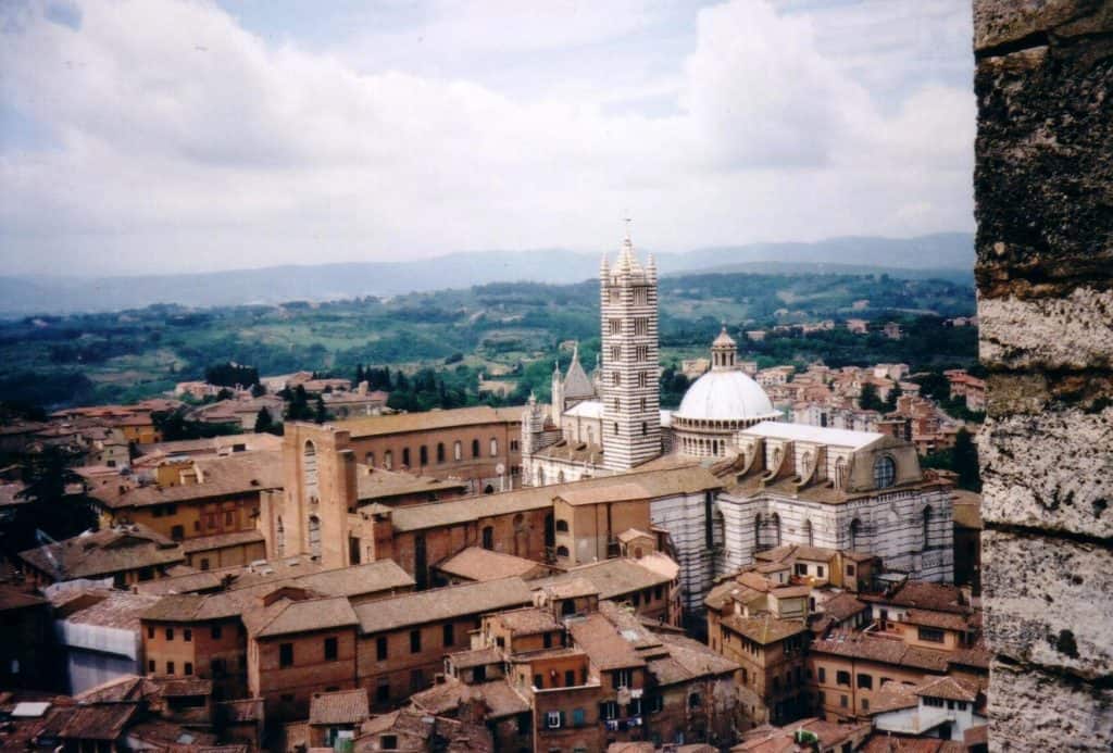 Elevated view of Siena from the City Hall tower, capturing the historic center with its medieval buildings and the famous white-and-brown striped bell tower and dome of the Siena Cathedral (Duomo di Siena), against a backdrop of the Tuscan countryside.