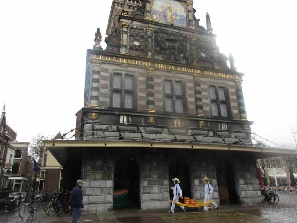 The historic Alkmaar Cheese Market building in the Netherlands, with cheese porters in traditional white uniforms and straw hats carrying cheese wheels. The ornate façade of the building, featuring intricate carvings and the Latin phrase, adds to the cultural significance of this renowned trading spot.