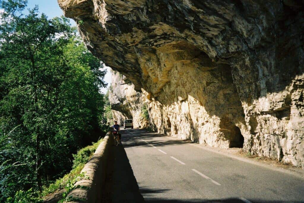 A cyclist rides along a road carved into the side of a cliff overlooking the River Lot in France, surrounded by lush greenery and the impressive overhang of rock formations.