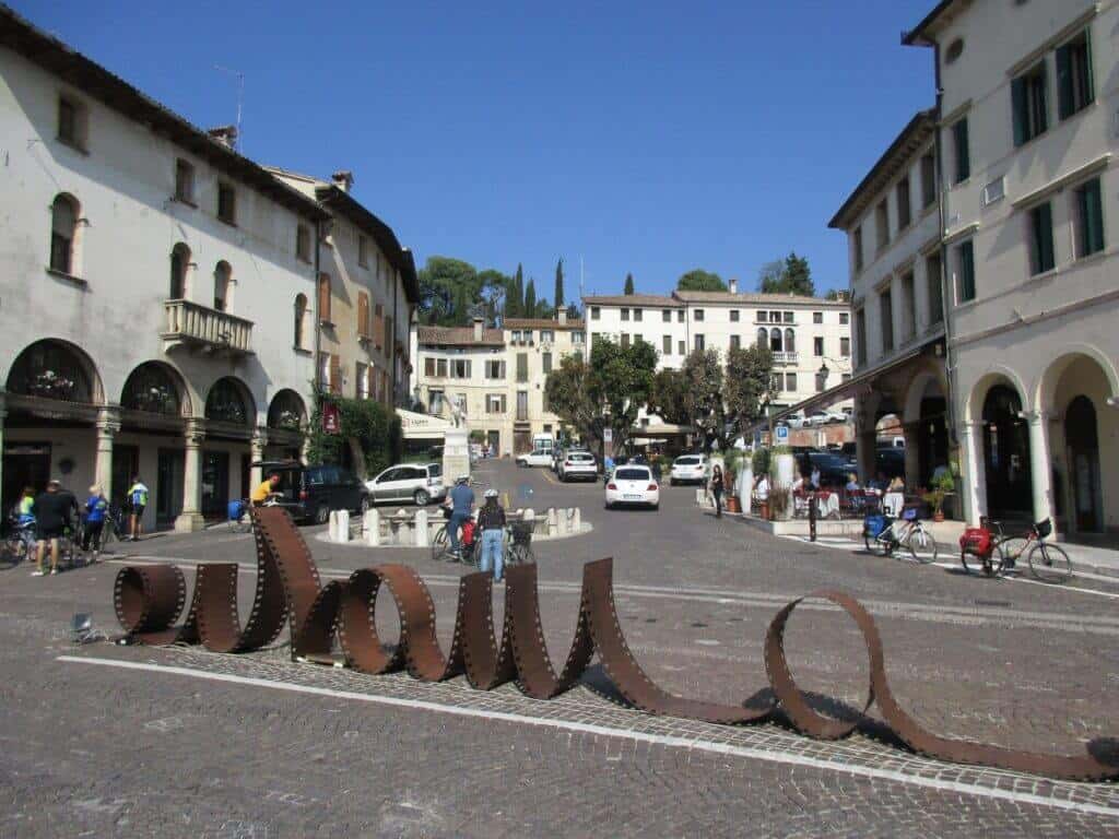 The bustling town square of Asolo, Italy, with pedestrians and cyclists near an artistic, rusted iron sculpture spelling 'Asolo', surrounded by historical buildings with arcades and a clear blue sky above.