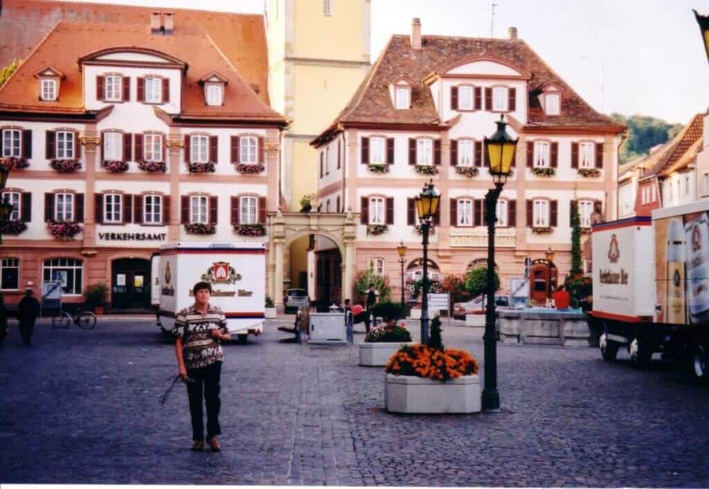 A vibrant scene from the main square in Bad Mergentheim, Germany, featuring traditional German architecture. The square is lined with charming buildings adorned with flower boxes under their windows, street lamps, and a central area with large flower planters. A delivery truck and a pedestrian add a dynamic element to this picturesque setting