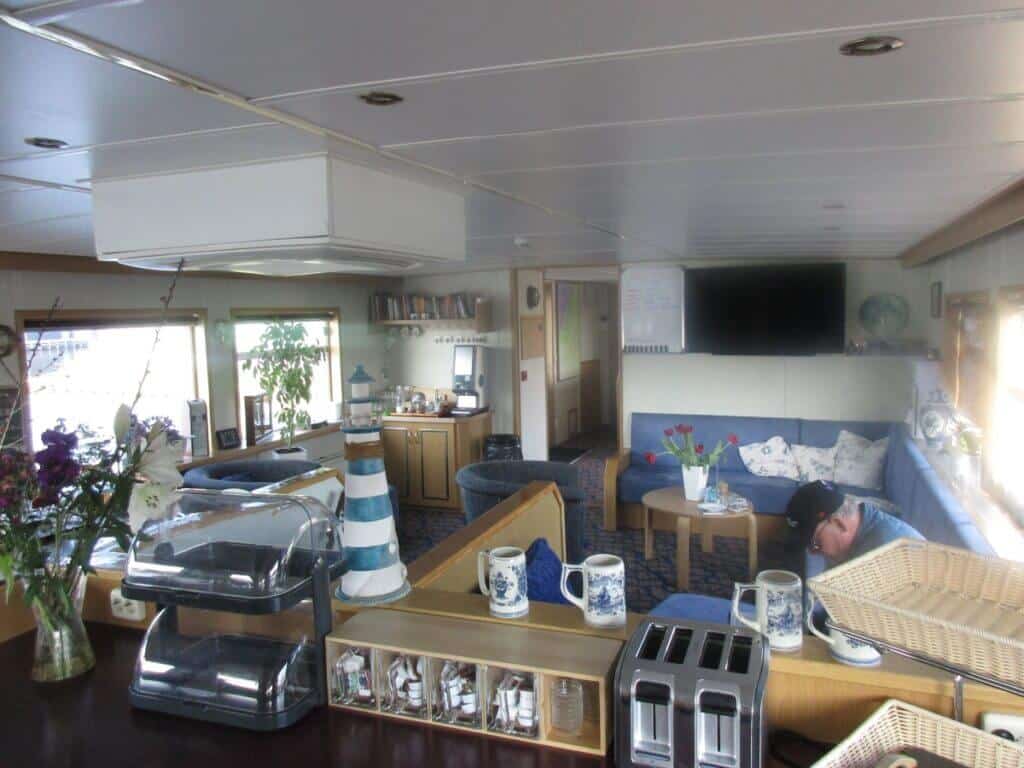 The cozy lounge area inside the 'Zwaantje' barge features comfortable seating, maritime-themed décor, and large windows letting in ample sunlight. The inviting space is well-prepared for guests, with coffee tables set with fresh flowers and neatly arranged dining settings, promising relaxation and camaraderie during the Holland Bike and Barge tour.