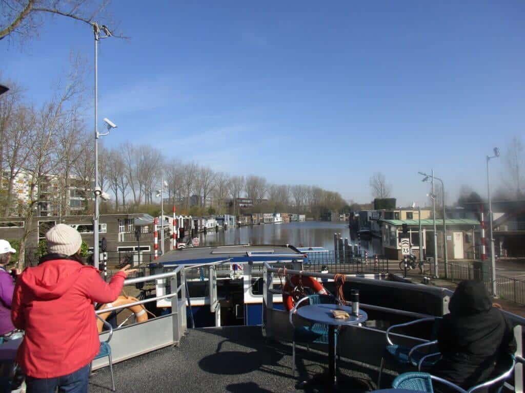 Passengers on a barge cruise along a canal leading to Amsterdam, with a view of leafless trees and residential houses along the waterway. The relaxed atmosphere on deck under the clear blue sky offers a unique perspective of approaching the vibrant city by water.