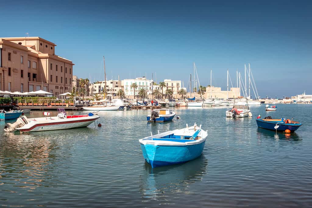A serene harbor scene in Bari, Puglia, featuring an array of boats gently floating on the calm water. Small boats, including one vivid blue rowboat, are moored close to the shore, while several yachts and sailing boats are anchored further out. The backdrop shows a line of waterfront buildings under a clear blue sky, enhancing the picturesque Mediterranean setting.