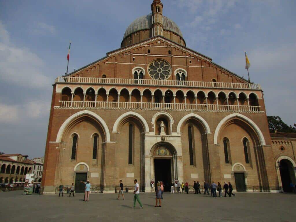 Visitors gather in front of the imposing Basilica di Sant'Antonio in Padua, Italy, an architectural masterpiece with a large domed roof and ornate arches, the Italian flag waving atop its faรงade under a partly cloudy sky.