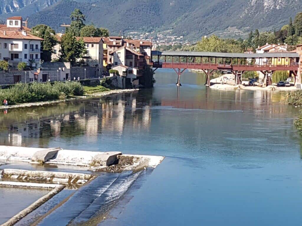 A serene view of the Brenta River in Bassano del Grappa, Italy, with the iconic Ponte degli Alpini, a covered wooden bridge, in the midground, traditional Italian houses along the riverbank, and the reflections on the calm water surface with a backdrop of green mountains.