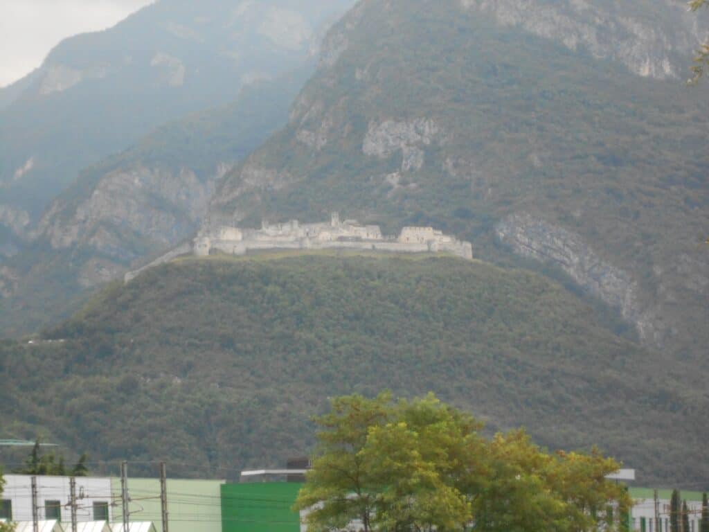 This image offers a distant view of Castel Beseno, a large medieval fortress perched atop a lush, wooded hill in northern Italy. The castle, enveloped in mist, dominates the landscape, its imposing walls and towers set against the backdrop of the rugged, forested mountains. The foreground features soft green treetops and a glimpse of industrial buildings