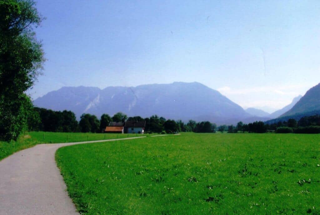 Scenic bike path near Bad Reichenhall, Germany, meandering through vibrant green fields towards the towering misty mountains, with a clear blue sky overhead