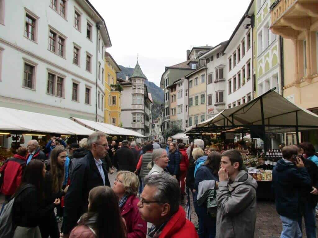 This image captures a lively scene at a market in Bolzano, Italy, bustling with activity and visitors. The narrow street, lined with colorful buildings of traditional architecture, is filled with market stalls under canopies and a crowd of people engaged in shopping and conversation. The atmosphere is vibrant and social, with people of various ages and appearances, contributing to the dynamic and cultural richness of the market day in Bolzano.