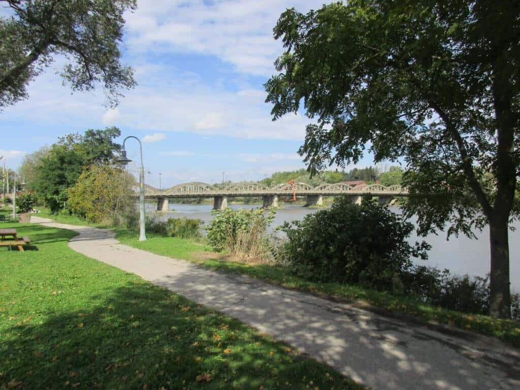 A serene view of the Grand River as it flows through Caledonia, Ontario. A picturesque pathway lined with lush trees and a park bench runs alongside the river, leading the eye towards a distinctive metal truss bridge in the distance. The bridge spans the calm, reflective waters of the river, set against a backdrop of clear skies and greenery