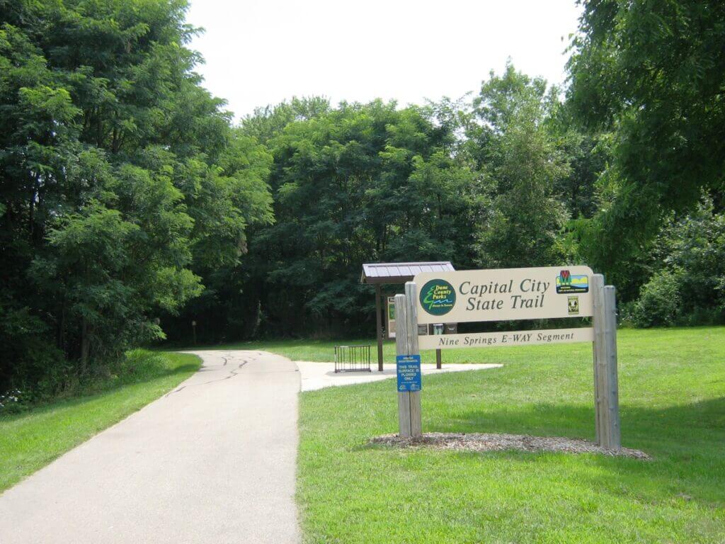 The entrance to the Capital City State Trail, marked by a wooden sign, located in a lush green park. The sign reads "Capital City State Trail, Nine Springs E-Way Segment" and includes logos for Dane County Parks, Friends of Capital Springs, and the Wisconsin Department of Natural Resources. The paved trail winds into a dense canopy of trees, inviting visitors to explore the serene natural surroundings.