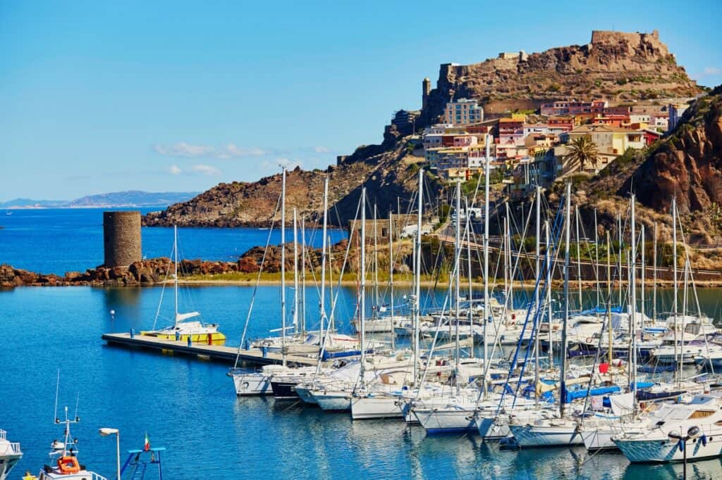A picturesque view of Castelsardo Village and its castle in Sardinia, Italy. The image captures a vibrant marina filled with an array of sailing boats in the foreground, with the azure Mediterranean Sea extending into the background. Above the marina, the historic village climbs up a rugged hill, topped by the imposing castle