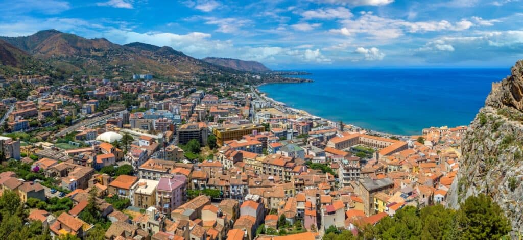 A panoramic view of Cefalù, a coastal town in Sicily, Italy, captured from a high vantage point. The photo showcases the dense clustering of traditional terracotta-roofed buildings stretching towards a vivid blue sea along the coastline. The backdrop features rolling hills and mountains that frame the town