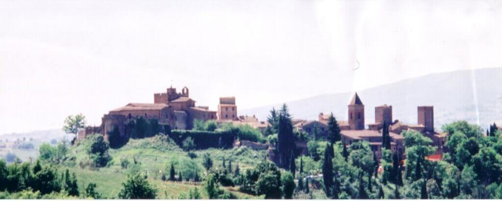 A distant view of the hilltop village of Certaldo, Italy, with its historic red-roofed buildings and fortifications set among green hills. The timeless architecture captures the essence of traditional Tuscan settlements, nestled in a peaceful countryside setting
