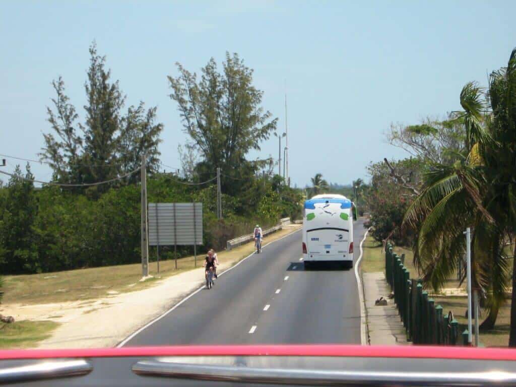 My Cuba Cycle Tour on a sunny day on a coastal road near Varadero, showing a cyclist and a tour bus traveling in opposite directions. The road is lined with lush tropical vegetation and palm trees, reflecting a serene, vacation-friendly atmosphere. The clear blue sky and the distant horizon enhance the tranquil setting.
