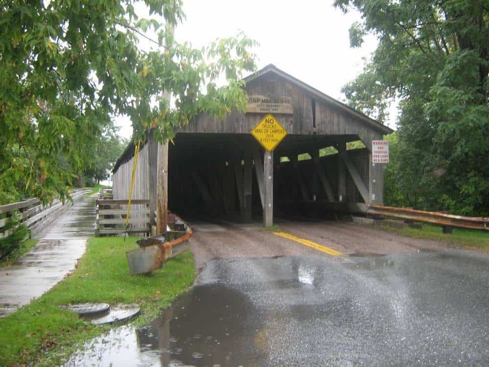 Historic wooden covered bridge on a rainy day in Middlebury, Vermont. The bridge features a weathered façade with warning signs about vehicle weight limits. Wet leaves cling to the branches of nearby trees, reflecting on the glistening wet road leading into the bridge