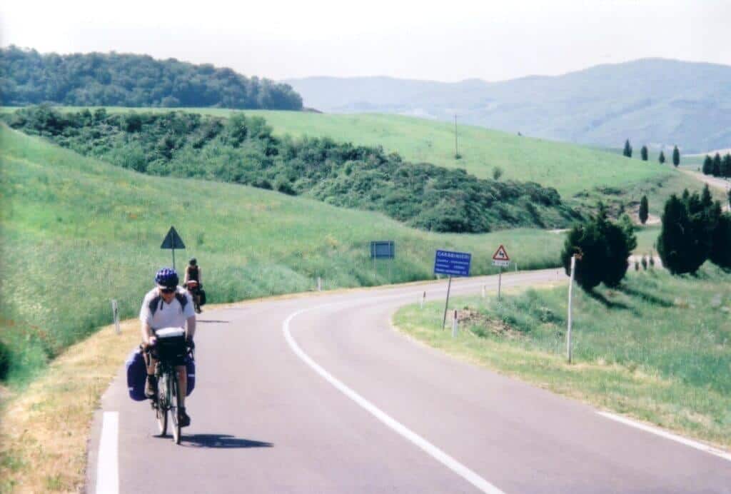 Cyclists on a tranquil road amidst the rolling green hills near Murlo, Tuscany, with road signs indicating a right-hand curve ahead. The idyllic Tuscan landscape stretches into the distance