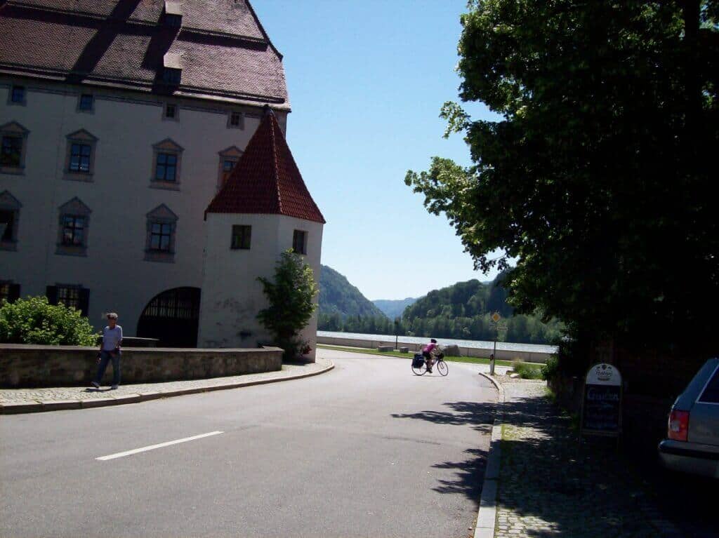 A cyclist rides along the Danube Cycling Route just after entering Austria, passing by an imposing traditional building and lush roadside trees, with the serene Danube river in the background.