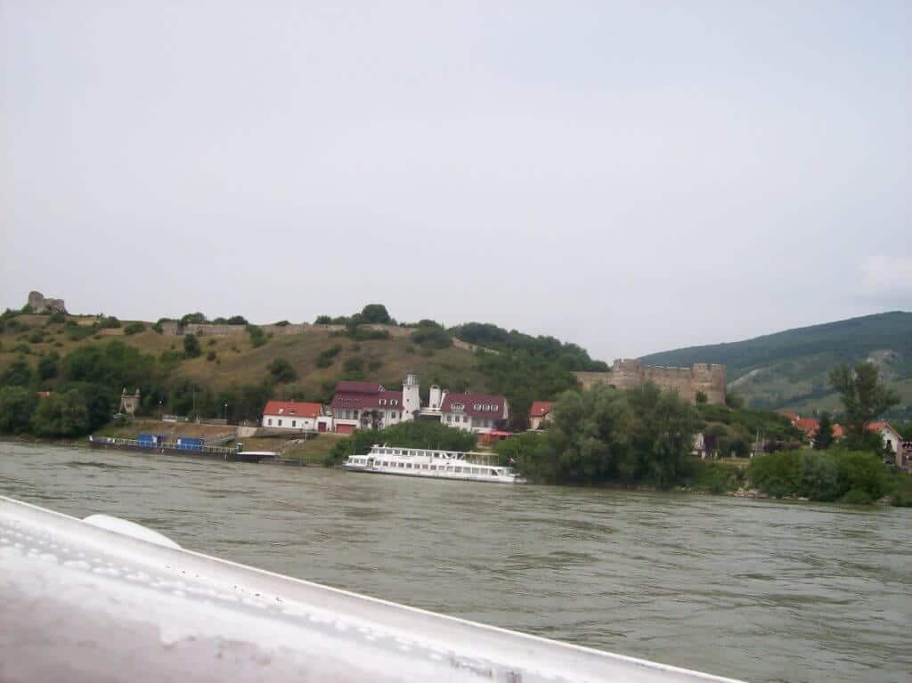 View from a boat showing the lush, hilly shoreline of the Danube River in Hungary. The scene includes a small white boat cruising close to the riverbank, a quaint village with red-roofed buildings, and ancient ruins on a hilltop, under a cloudy sky.