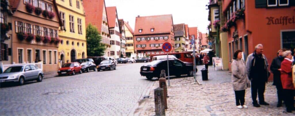 A bustling cobblestone street in Dinkelsbรผhl, Germany, lined with colorful, traditional German buildings adorned with flower boxes. The street scene is lively with parked cars and pedestrians engaging in conversation, showcasing the charming, vibrant atmosphere of this historic town.