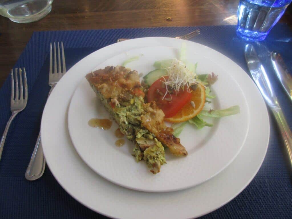 A plated dinner on the 'Zwaantje' barge, featuring a savory slice of quiche accompanied by a fresh salad with sprouts, cucumber, and citrus. The simple yet elegant presentation on white tableware, set against a blue placemat, offers a taste of the culinary experiences provided on the bike and barge tour.