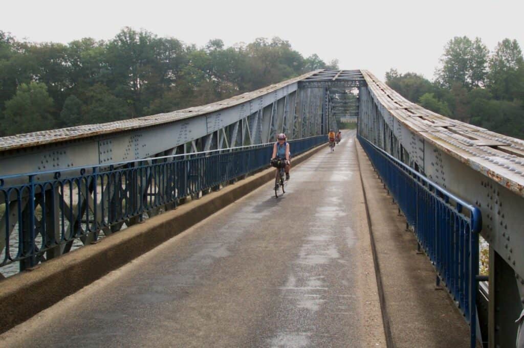 Cyclists cross an old metal truss bridge over the Dordogne River in France, the structure's blue railings and weathered steel creating a striking contrast against the surrounding greenery on a hazy day.