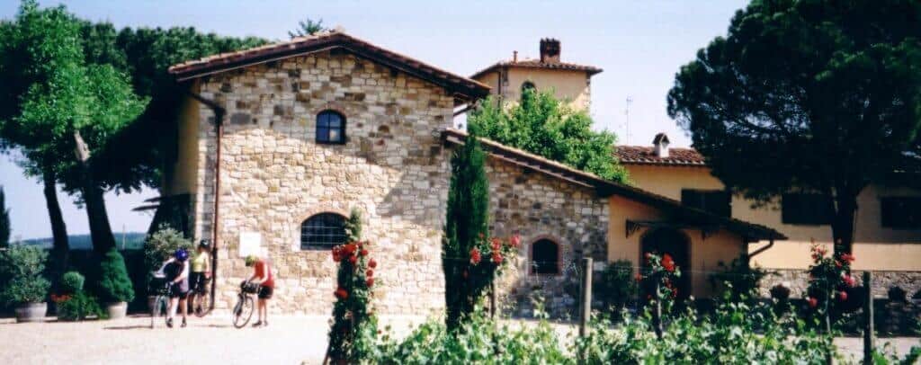 Cyclists pause at the charming Fattoria Viticcio winery in Tuscany, framed by stone buildings with terracotta roofs and flourishing red roses. The estate's greenery and tranquil setting invite visitors to explore the delights of Tuscan winemaking