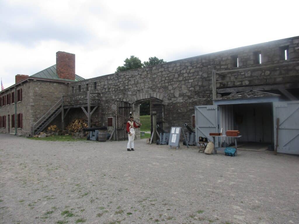 Fort Erie in Ontario, showcasing the fort's robust stone structures with wooden accents. A person dressed in traditional military attire from the 1800s stands guard, adding a touch of authenticity and historical context to the scene. The open courtyard area, where various historical artifacts and informational signs are displayed, invites visitors to explore and learn more about the fort's historical significance.