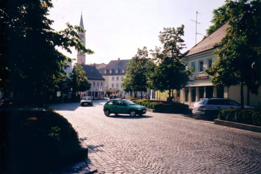 A tranquil morning in the town square of Friedberg, Germany, captured in a photograph showing a cobblestone street lined with classic European architecture. A few cars are parked along the street, and the peaceful ambiance is accentuated by mature trees casting gentle shadows. The town's spire stands elegantly in the background, adding to the historic charm of the scene.