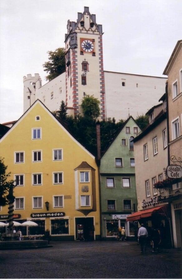 Street-level view of colorful buildings in Füssen, Germany, with a focus on the historic clock tower of Füssen Castle rising above, featuring its iconic red and white exterior against a backdrop of overcast sky.