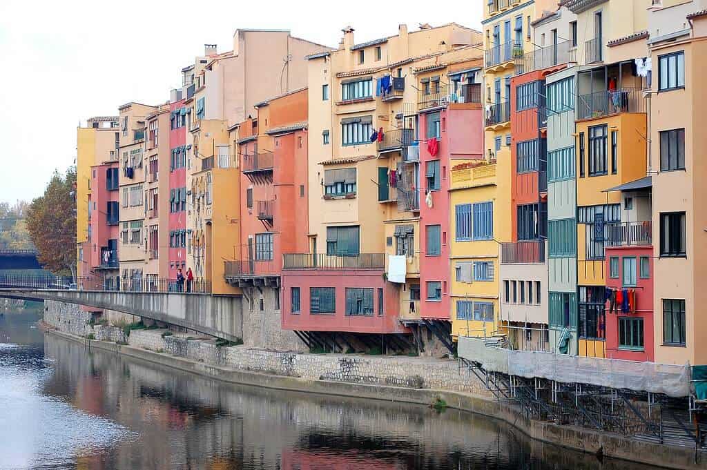 Colorful houses with vibrant facades line the Onyar River in Girona, Spain. The image shows the picturesque riverside architecture with reflections in the water, characteristic of the old city's charm and cultural heritage.