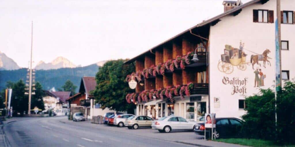 The Gasthof Zur Post in Schwangau, Germany, captured at twilight with the majestic Alps in the background. This traditional Bavarian guesthouse is adorned with vibrant flower boxes and features a large mural depicting a historic carriage scene.