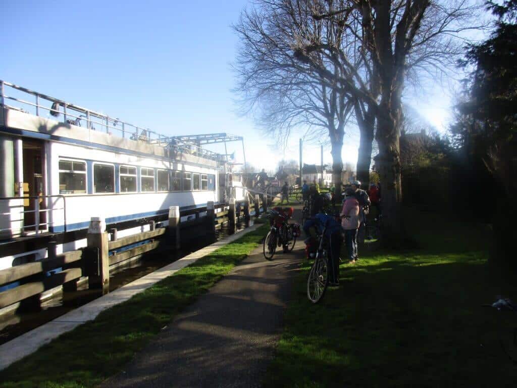 Cyclists gather beside a barge on a sunlit path in Holland, preparing for a day of exploration on a Bike and Barge tour. The tall, leafless trees and the barge's reflection on the canal water create a sense of anticipation for the journey ahead in this Dutch setting.