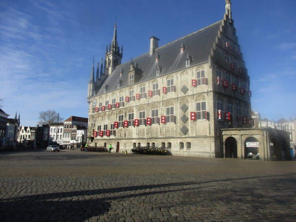 The historic Market Square in Gouda basks in the sunlight, showcasing the grandeur of the Gothic-style town hall with its striking red window shutters. The cobblestone square and a glimpse of the surrounding Dutch architecture invite exploration and admiration of this well-preserved cultural site
