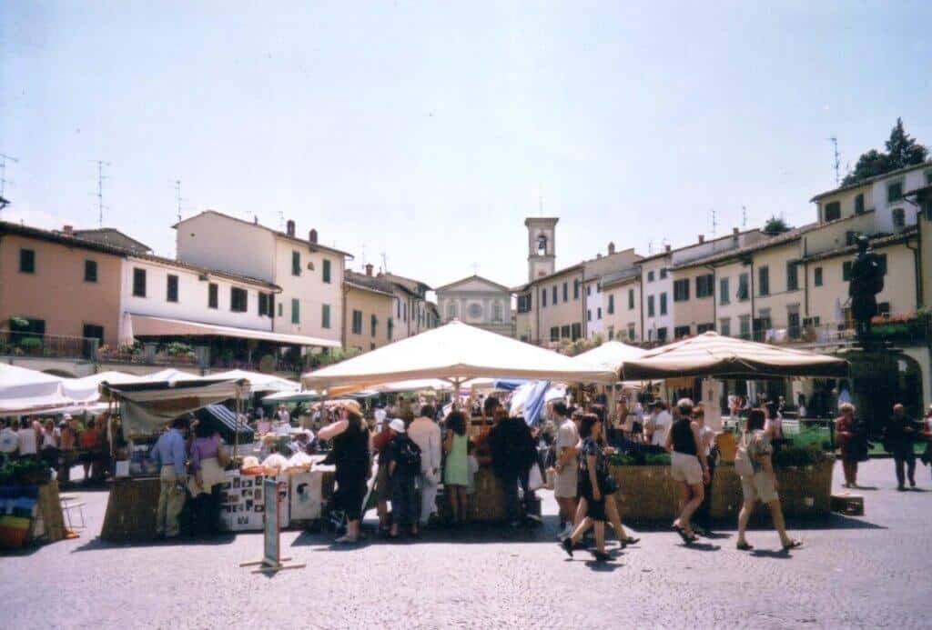 Busy market day in the sunlit square of Greve, Italy, with locals and tourists browsing stalls under white canopies offering a variety of goods. The surrounding buildings and a clock tower provide a quaint backdrop