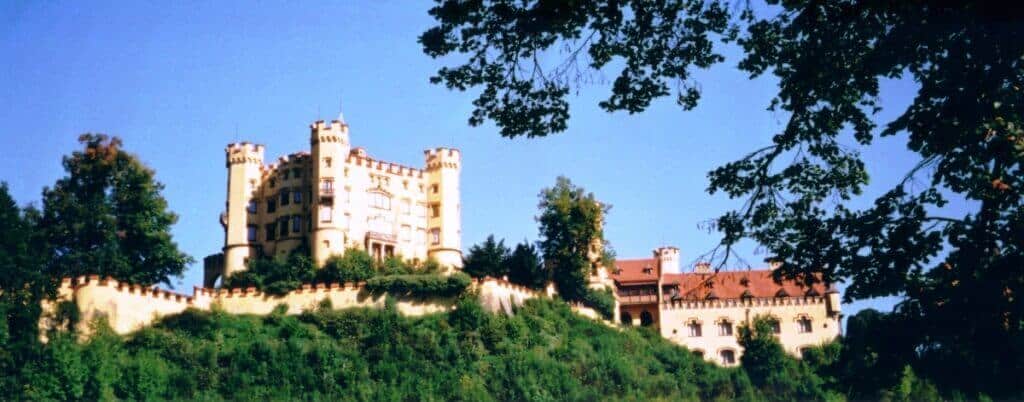 The historic Hohenschwangau Castle in Germany stands proudly on a hill, surrounded by verdant greenery and a clear blue sky, with the foreground framed by tree branches, invoking a fairytale ambiance.