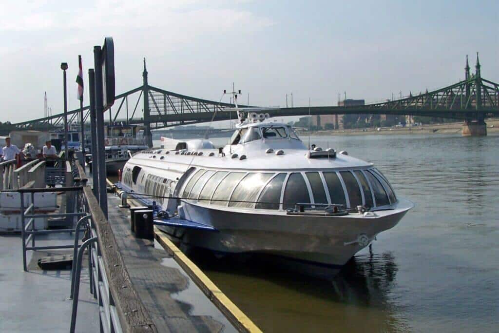A sleek hydrofoil boat moored at a dock on the Danube River in Budapest, with the iconic Liberty Bridge in the background. The boat, with its metallic finish and futuristic design, is ready to depart, while pedestrians and crew members prepare for the voyage on a sunny day