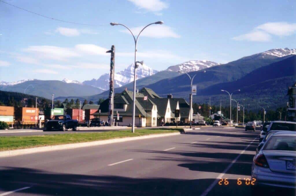View of the main street in Jasper, Alberta, showing a vibrant street scene with cars, a totem pole, and quaint architecture against a backdrop of the stunning Canadian Rockies.
