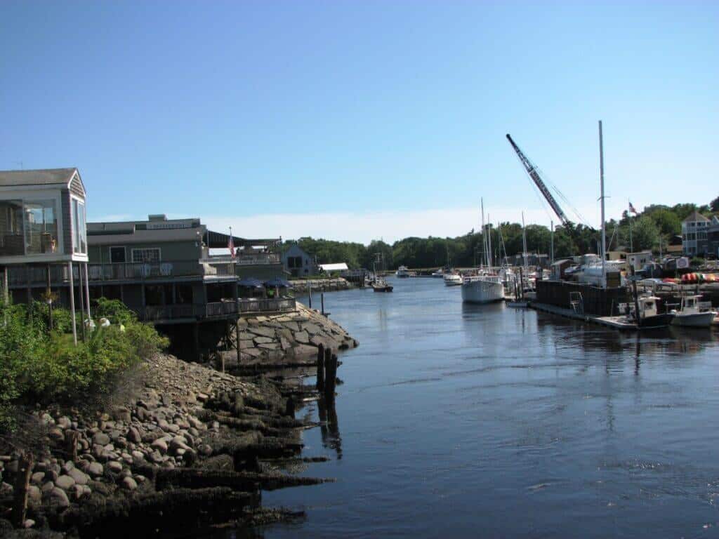 Serene river scene in Kennebunkport, Maine, with waterfront buildings, docks, and moored sailboats, under a clear blue sky, reflecting the town's maritime heritage and the leisurely pace of coastal life.