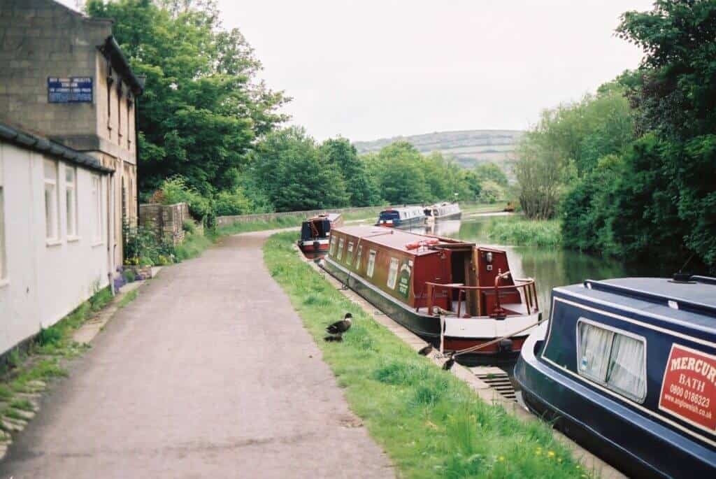 Narrowboats moored along the peaceful Kennet & Avon Canal in England, with a towpath inviting a leisurely stroll beside the waterway. The scene is framed by lush greenery and a hint of rolling hills in the distance, capturing the essence of the English countryside.