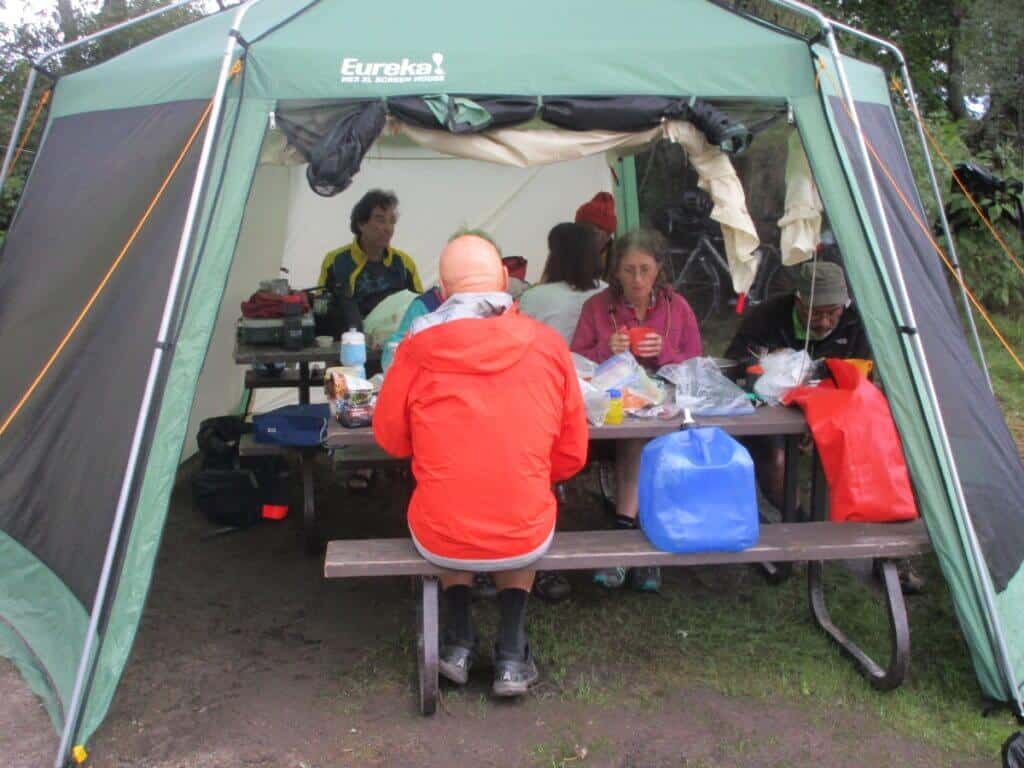 A group of campers gathered inside a large Eureka kitchen tent at a campsite. The tent is equipped with a picnic table where campers are engaged in meal preparation and socializing. Visible are various camping supplies like stoves, food containers, and water jugs, adding to the practical and communal atmosphere. The surrounding greenery hints at a secluded outdoor setting