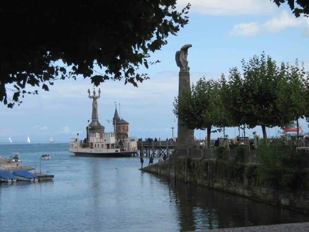 A serene view of Konstanz harbor in Germany, featuring a striking statue of Imperia at the harbor's entrance. The statue dominates the foreground, with sailboats and the blue waters of Lake Constance stretching into the background. Architectural features such as a historic pier building with twin turrets frame the scene, all under the canopy of lush green trees, adding depth and cultural richness to the waterfront setting.