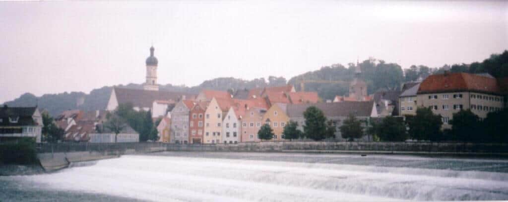 A misty view of Landsberg am Lech, Germany, showcasing a charming riverside scene with cascading waters of the Lech River. Colorful, traditional houses and prominent church spires create a picturesque skyline against a backdrop of gentle hills. The tranquil river and quaint architecture convey a peaceful, historic atmosphere in this old Bavarian town.