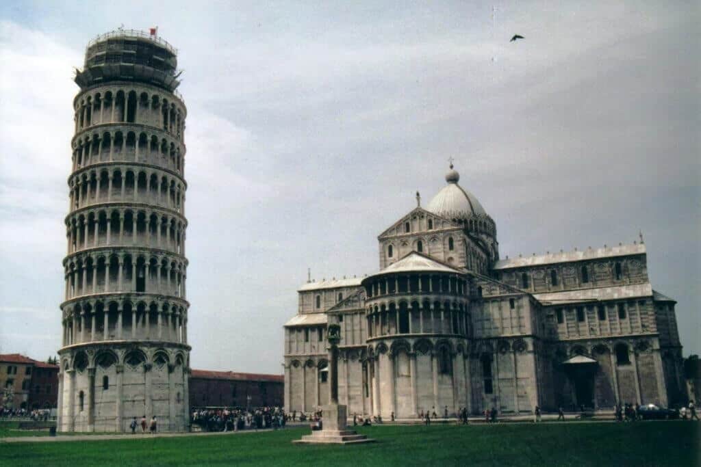 The Leaning Tower of Pisa, a freestanding bell tower, tilts noticeably beside the Pisa Cathedral, both showcasing exquisite Romanesque architecture. The Piazza del Duomo is dotted with visitors, under a partly cloudy sky, illustrating this iconic Italian landmark's enduring appeal.