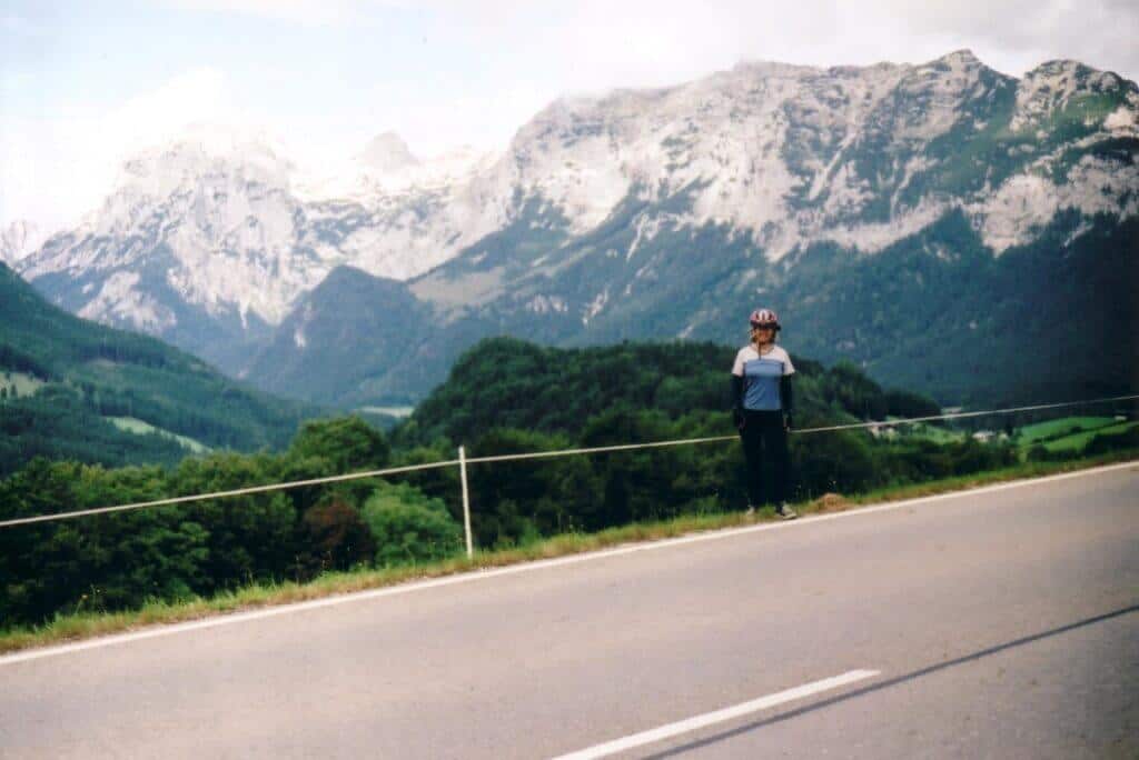 A cyclist stands beside a road leaving Berchtesgaden, framed by the majestic Alps in the background. The rugged peaks, still capped with snow, tower over lush green valleys, presenting a striking contrast. The cyclist, equipped with a helmet and casual attire, pauses to admire the dramatic landscape along this scenic route.
