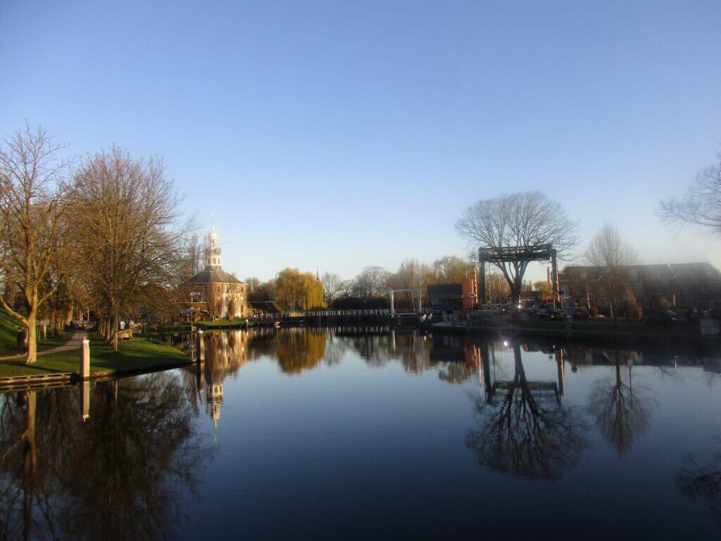 Early morning in Leiden, Netherlands, with the serene reflection of a historic church and a traditional drawbridge on the smooth surface of a canal. Leafless trees along the bank and the calm water create a tranquil and picturesque Dutch townscape.