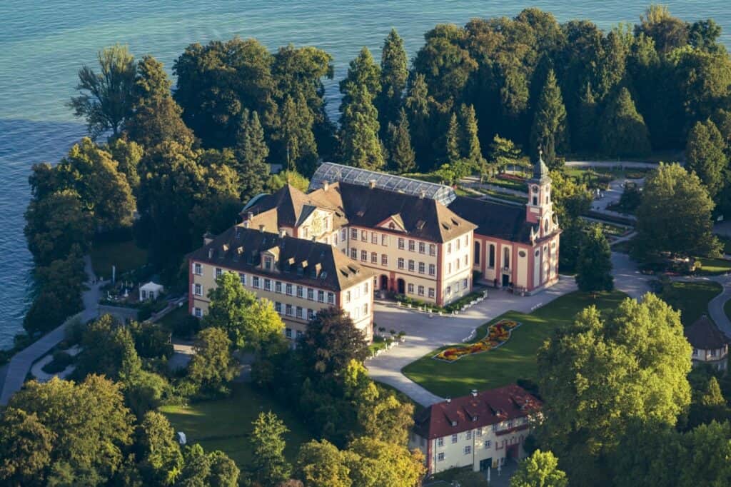 An aerial view of the Baroque palace located on Mainau Island, Germany, nestled amidst lush gardens and surrounded by the expansive Lake Constance. The palace, with its classic red and white facade, is complemented by an ornate church and a large greenhouse. The manicured gardens feature intricate floral designs and pathways